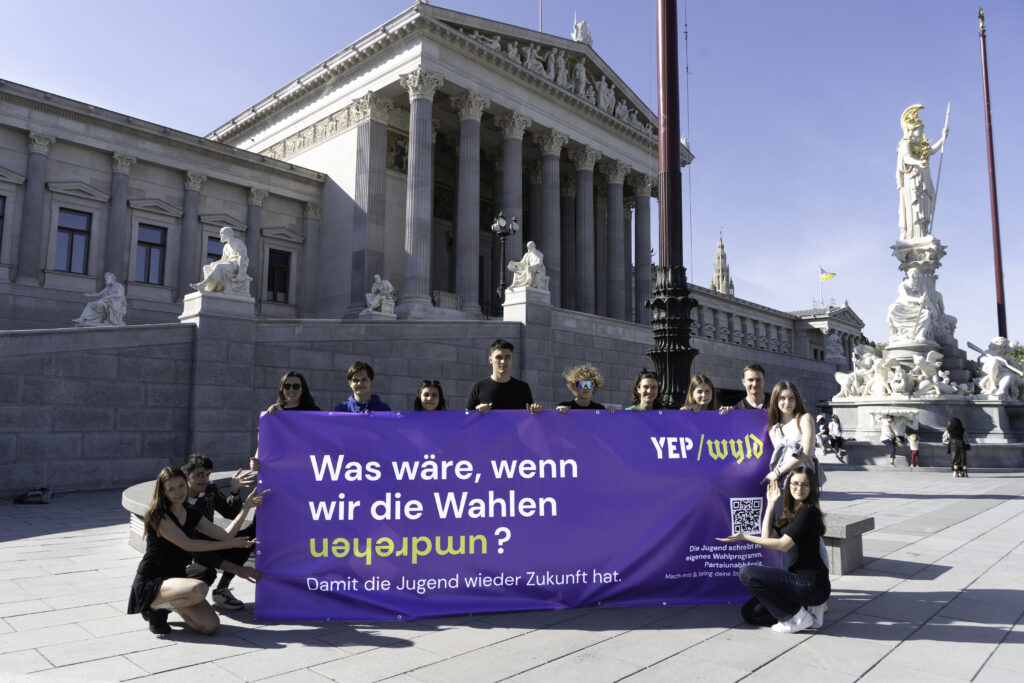 Eine Gruppe Jugendlicher steht vor dem Parlamentsgebäude in Wien und hält ein Transparent auf dem steht: Was wäre, wenn wir die Wahlen umdrehen? Das Wahlprogramm der unabhängigen Jugend.